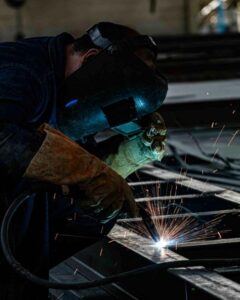 A person in a welding helmet and brown groves welding two pieces of metal together